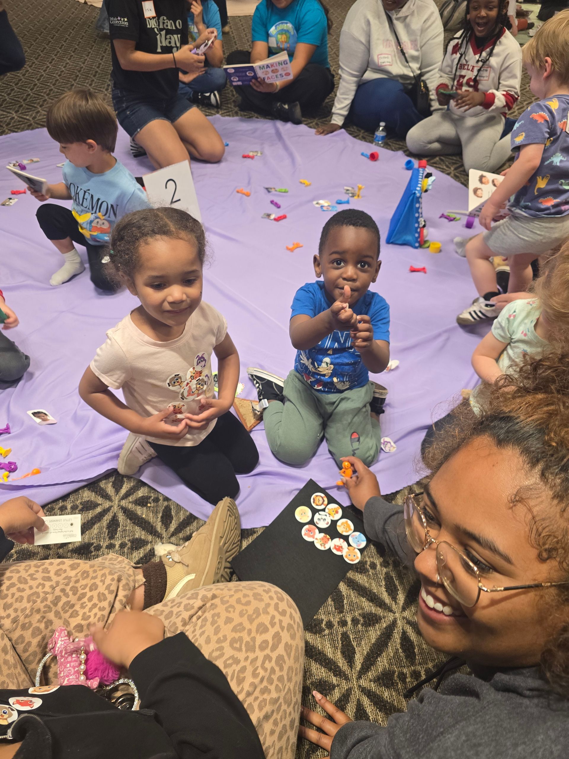 Children gathered on a purple mat, making crafts and snacks with adults nearby in a playful indoor setting