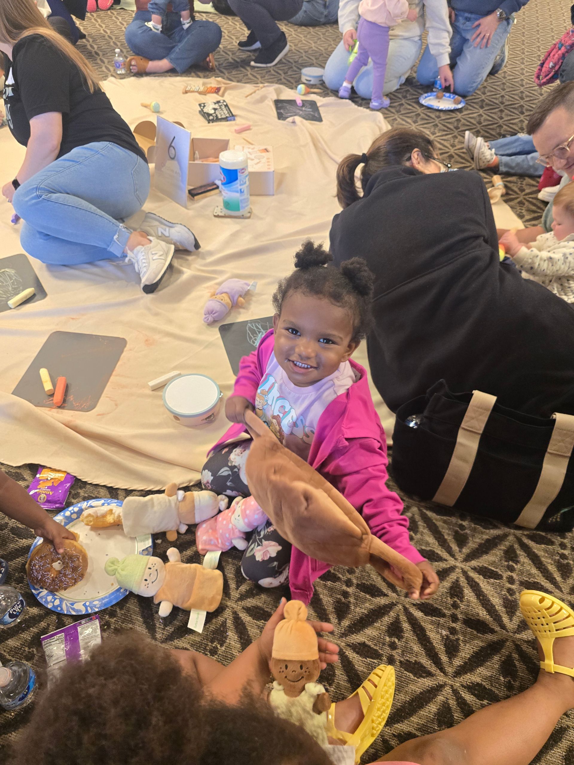 Children sitting on a floor craft area with supplies, one child in a pink jacket holding a stuffed animal and smiling.