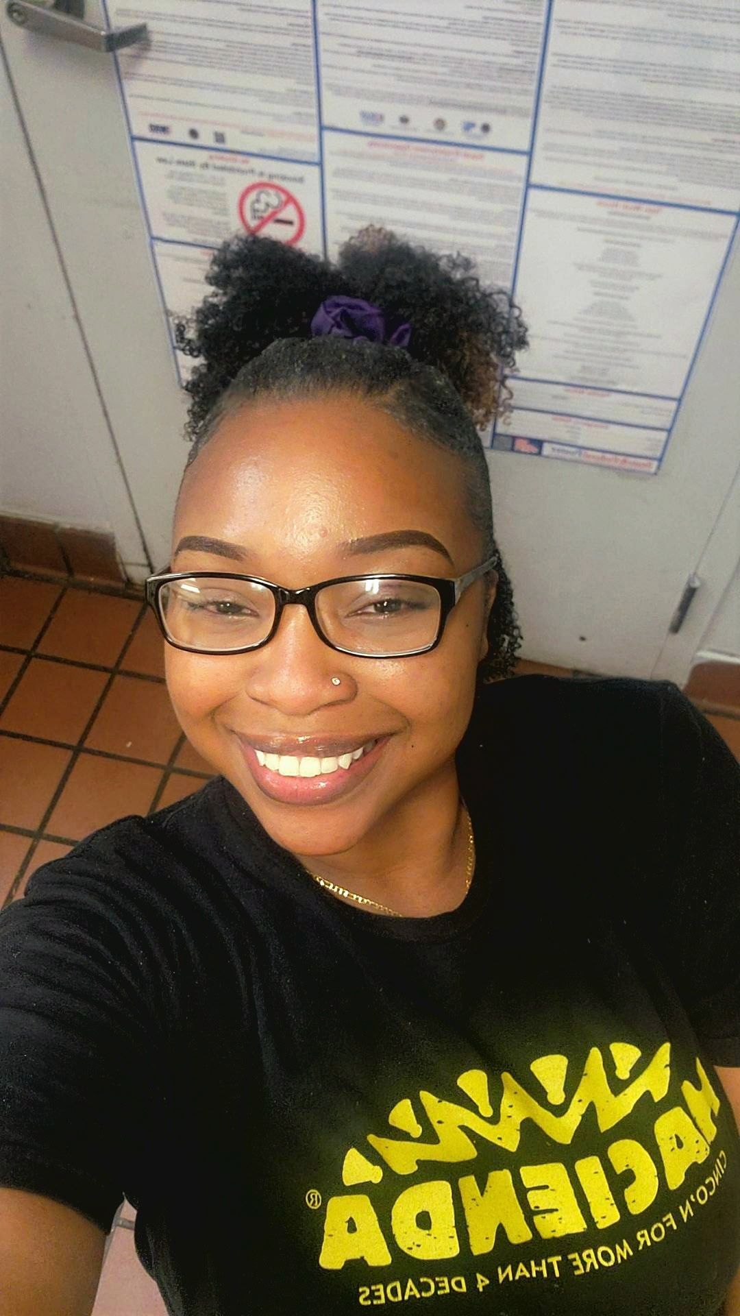 Smiling person taking a selfie in an office, wearing glasses and a black shirt with yellow text.