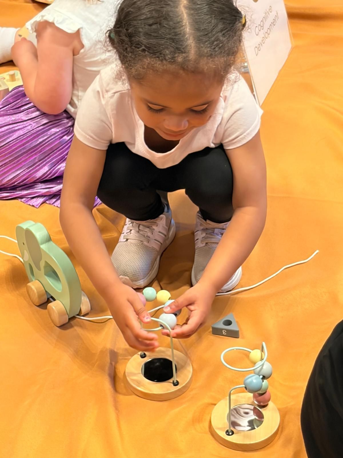 Child crouching on an orange floor, arranging beads over a wooden toy with another child nearby.