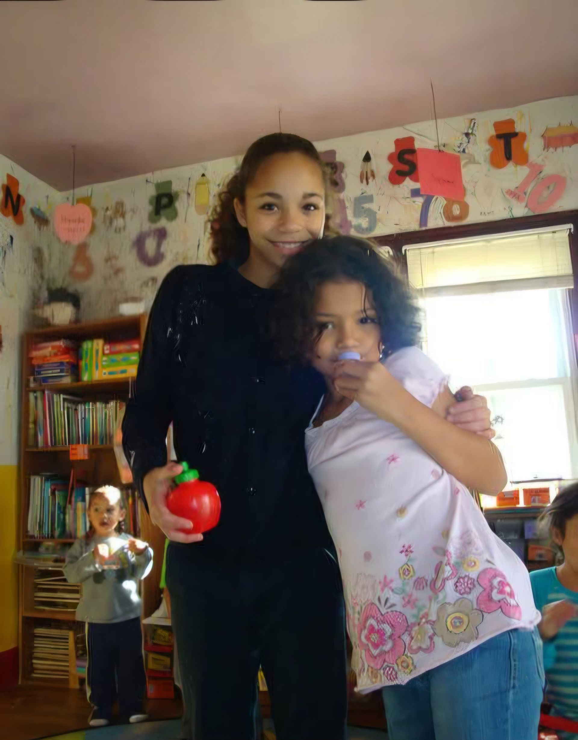 Two children posing in a classroom; one holds a red apple, with bookshelves and wall decorations behind them.