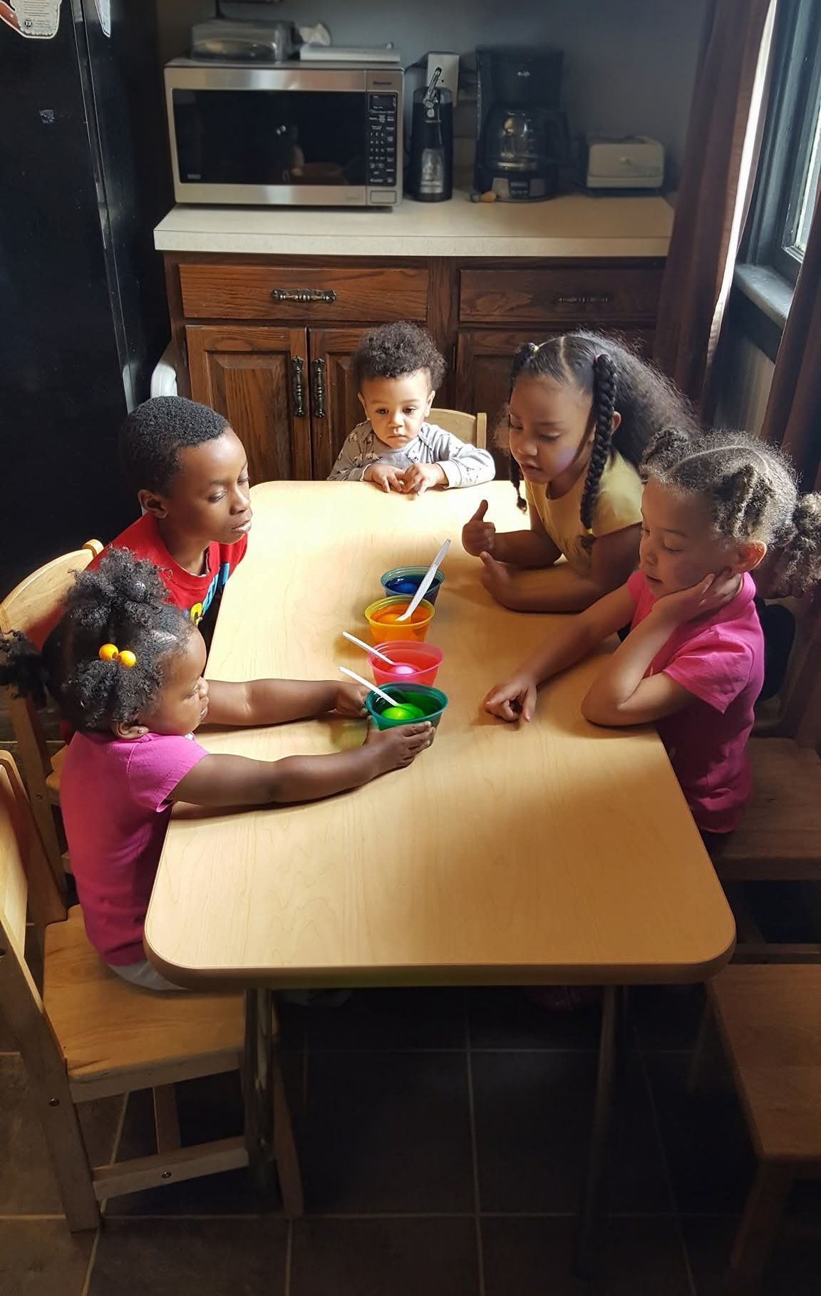 Four children sit around a kitchen table playing with colorful toys and cups.