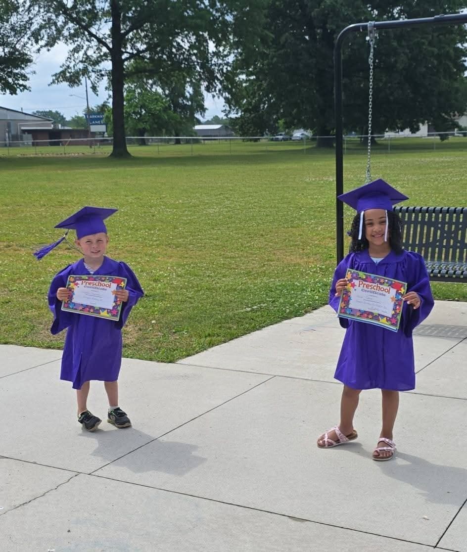 Two children in purple graduation caps and gowns holding certificates outdoors on a sidewalk.
