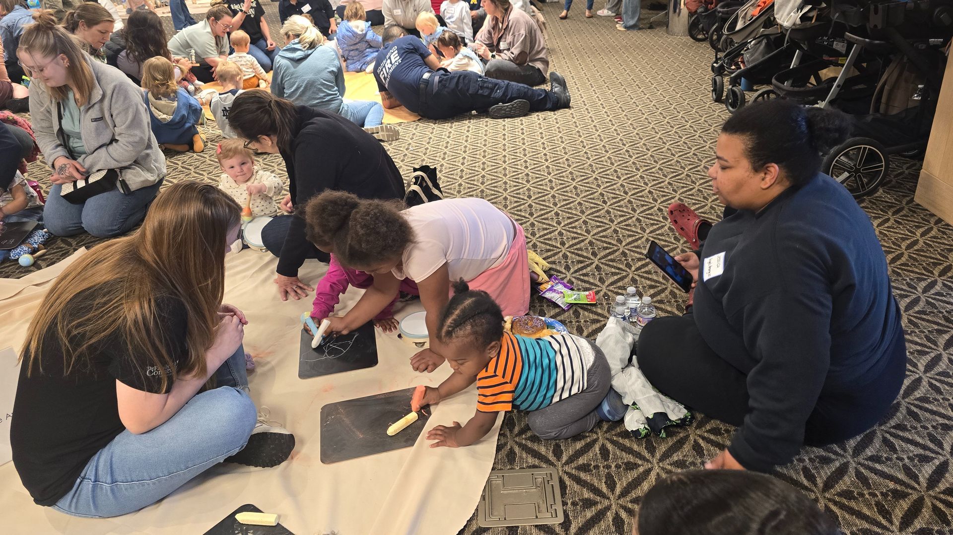 Children gathered on a carpeted floor, sitting and playing with colorful craft materials in a classroom-like space