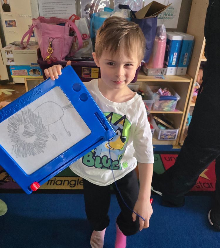 Child holding up a blue-framed drawing board in a classroom, smiling.