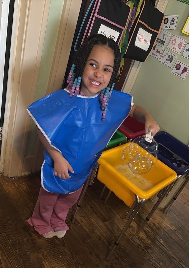 Smiling child wearing a blue bib stands by a yellow tray and holds a clear container in a classroom