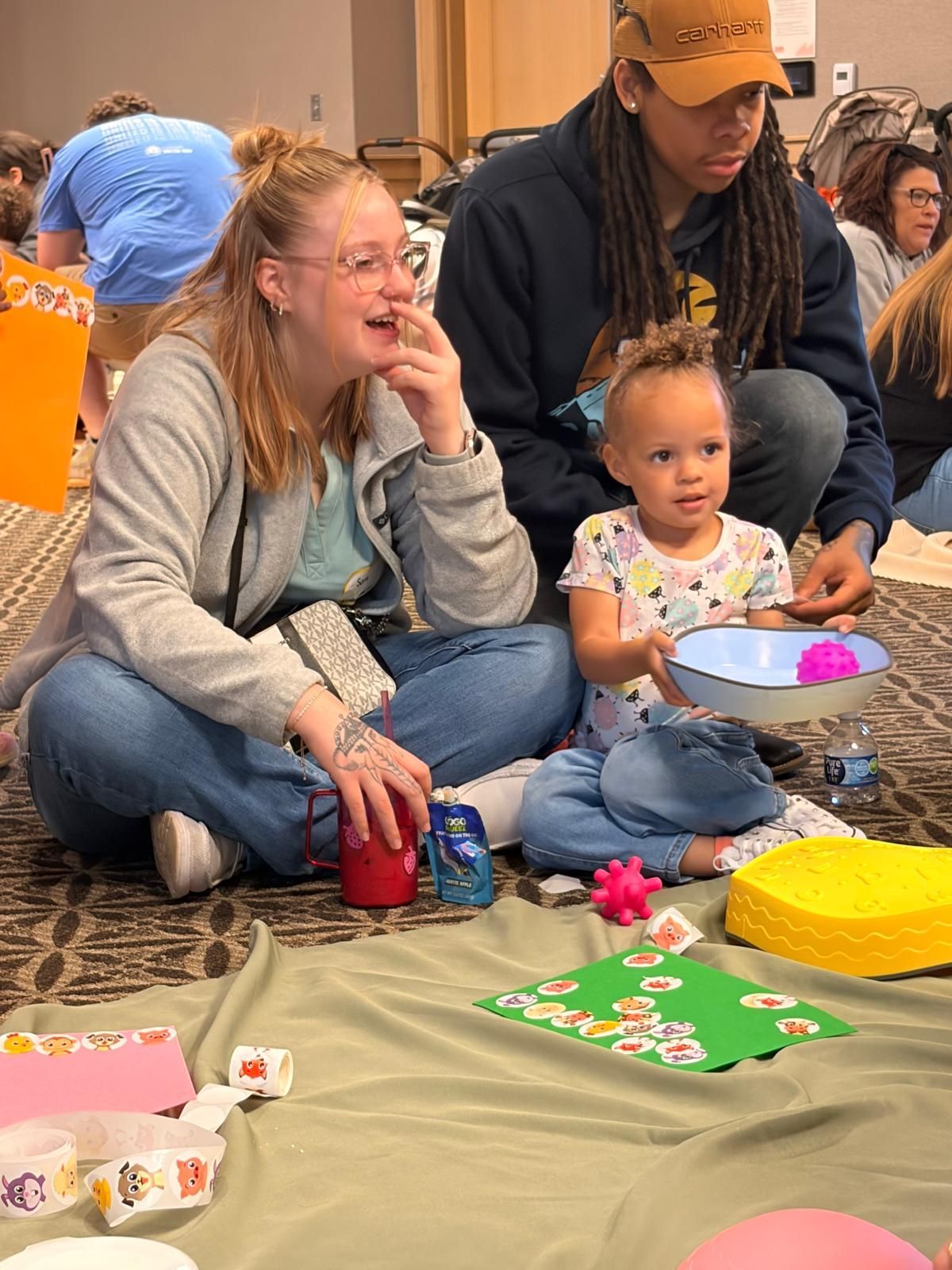People sitting on a floor at a family gathering, with a child holding a bowl and colorful items nearby.