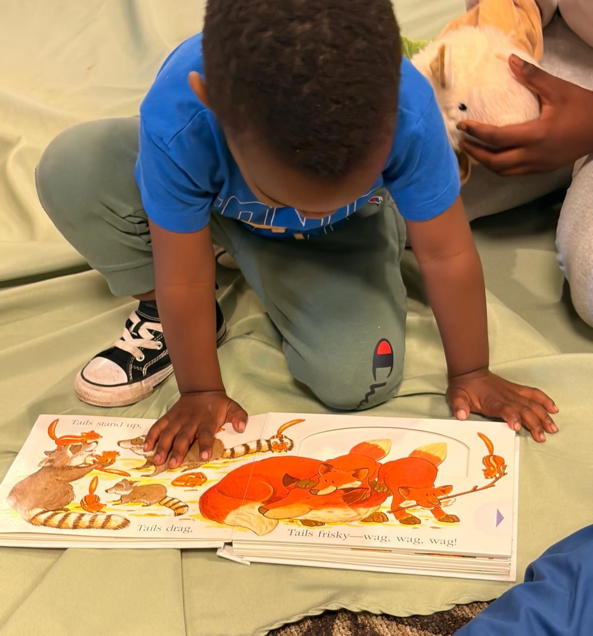 Child kneels over a colorful dinosaur book, touching the pages while an adult holds the book open.