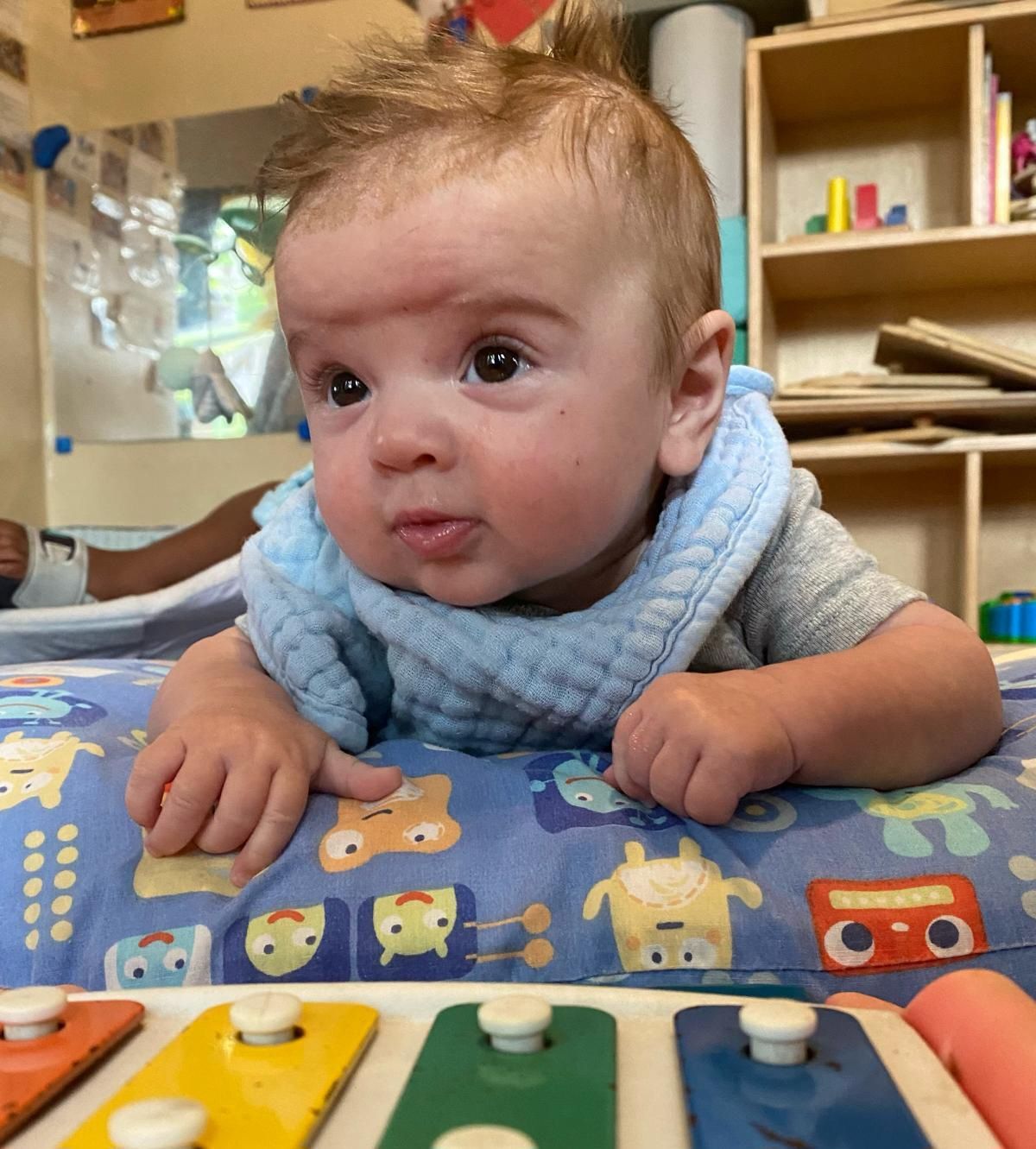 Baby leaning on a colorful play mat, looking up in a classroom or playroom.