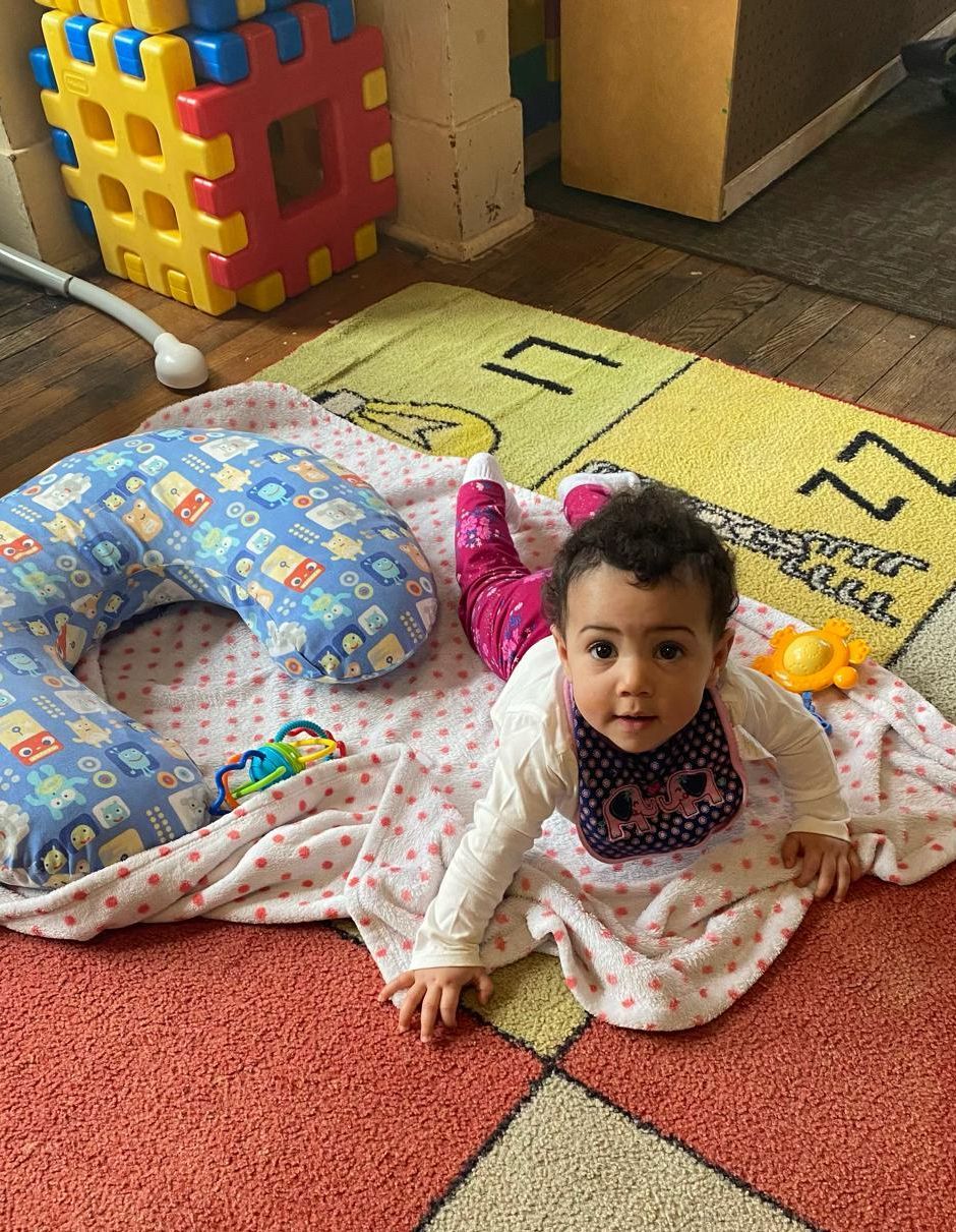 Baby sitting on a patterned play mat with toys and a blue support pillow in a colorful indoor play area