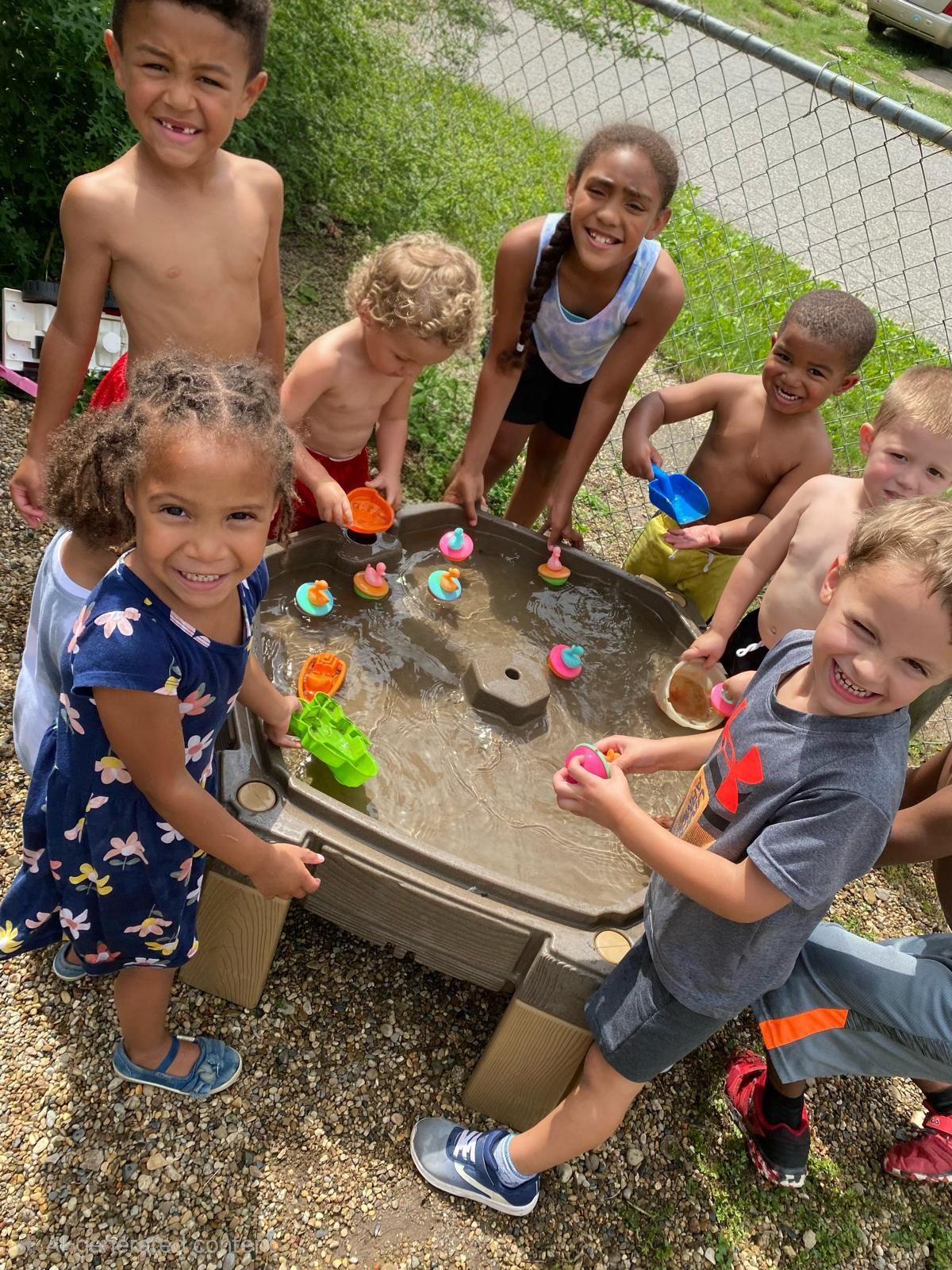 Children playing around a water table outdoors, smiling and splashing with colorful toys