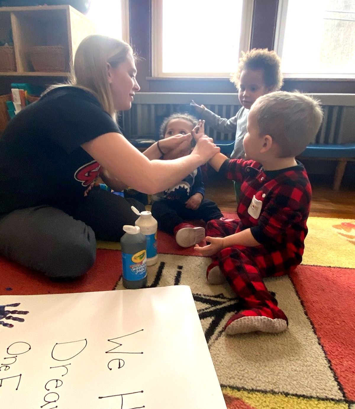 Adult helping two children sit on a rug, with a white board and toys in a bright room.