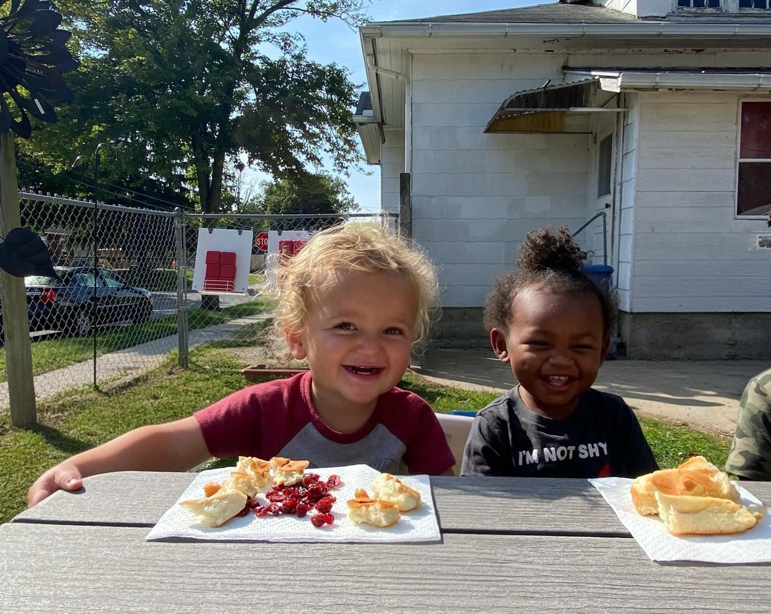 Two children smiling at a picnic table with plates of food in a sunny backyard