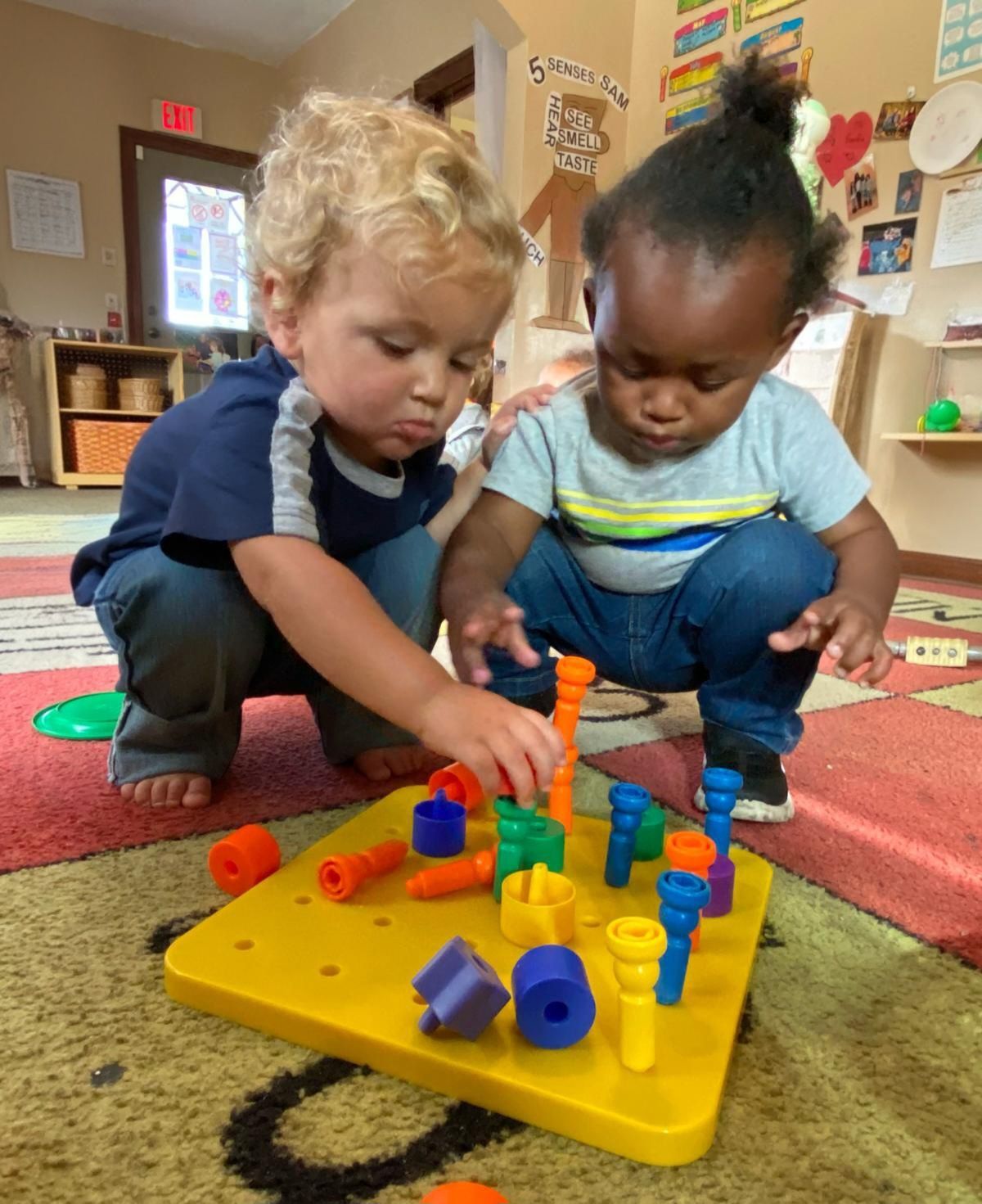 Two children crouch over a yellow block board in a classroom, building with colorful pegs.