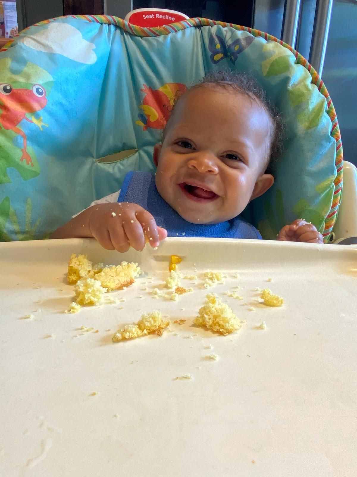 Smiling baby in a colorful high chair reaching for food on a white tray