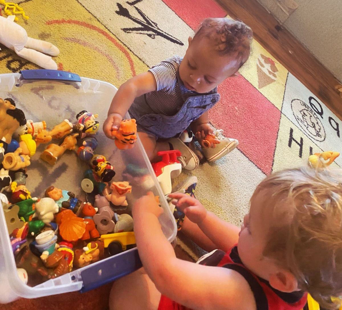 Two toddlers playing with toy animals beside a clear plastic bin on a colorful rug