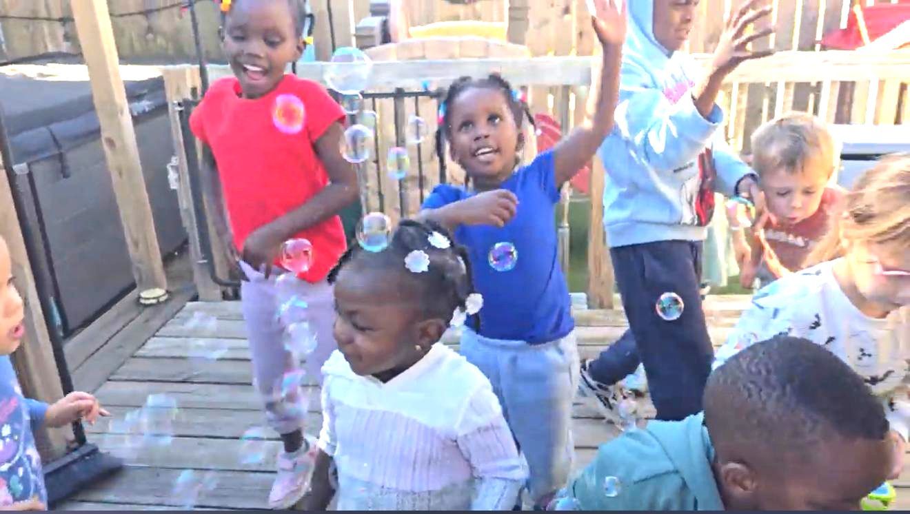 Children dancing and smiling on a wooden deck outdoors, with one child in a red shirt in front.