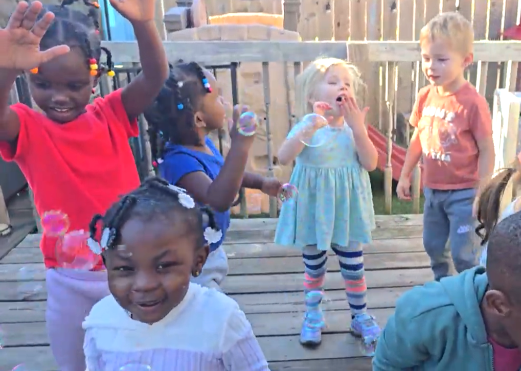 Children dancing and playing on a wooden deck, with one child in a red shirt raising both hands.