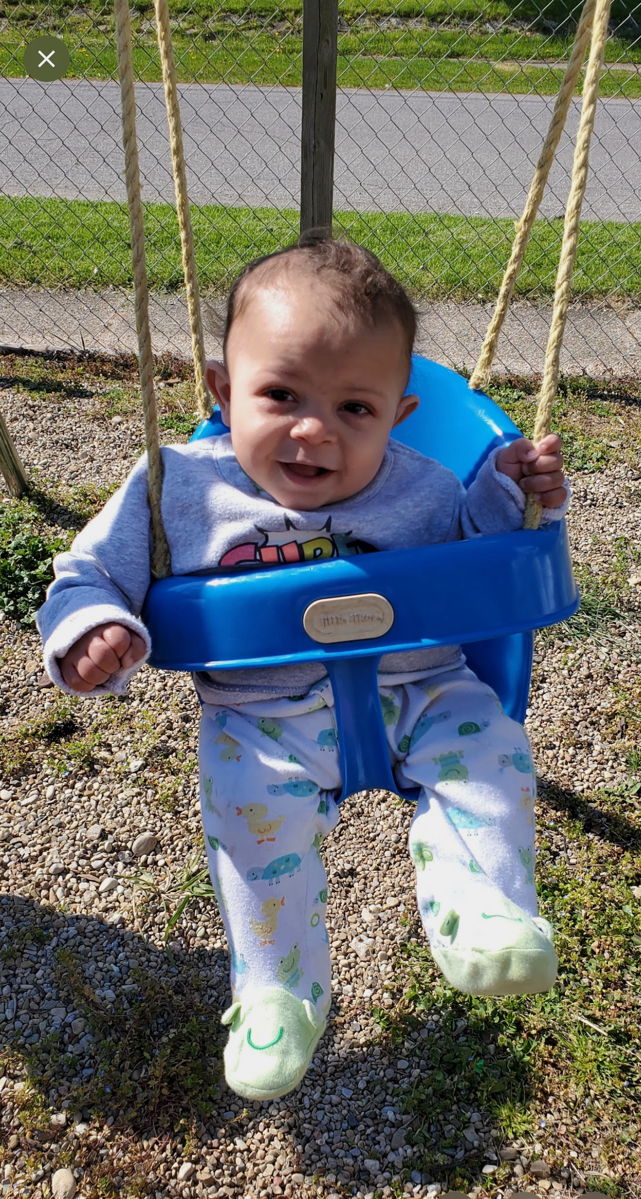 Baby in a blue swing outdoors, smiling with feet extended over gravel.