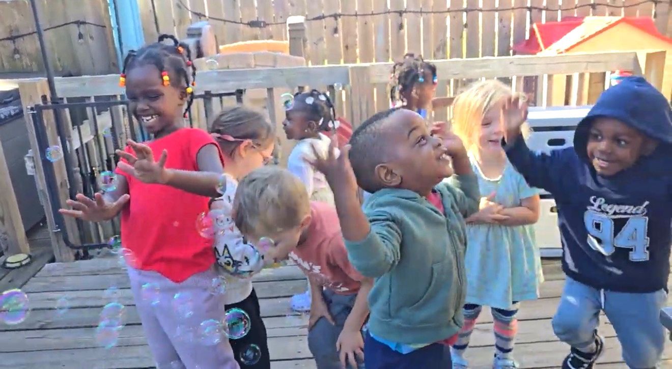 Children dancing and laughing outdoors on a wooden deck in colorful clothes.