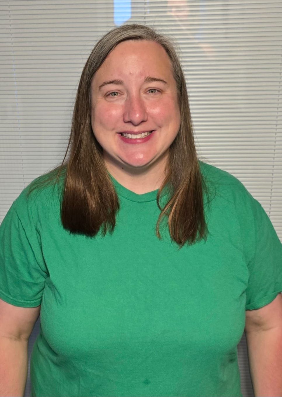Smiling person in a green shirt standing in front of window blinds.