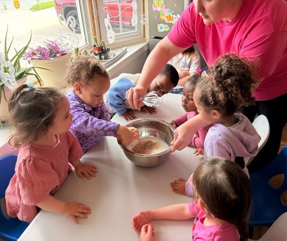 Children around a table mixing batter in a bowl with an adult nearby, in a bright classroom setting