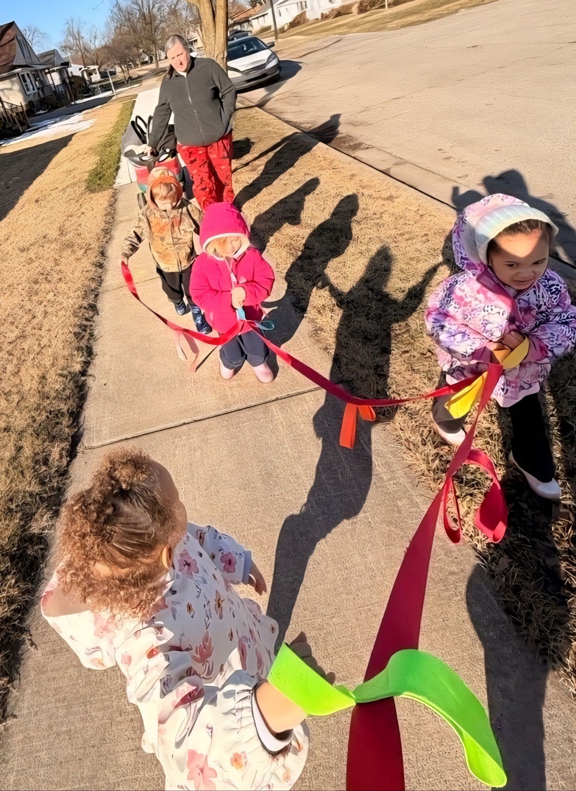 Children holding a colorful ribbon on a sidewalk, with an adult nearby on a sunny day.
