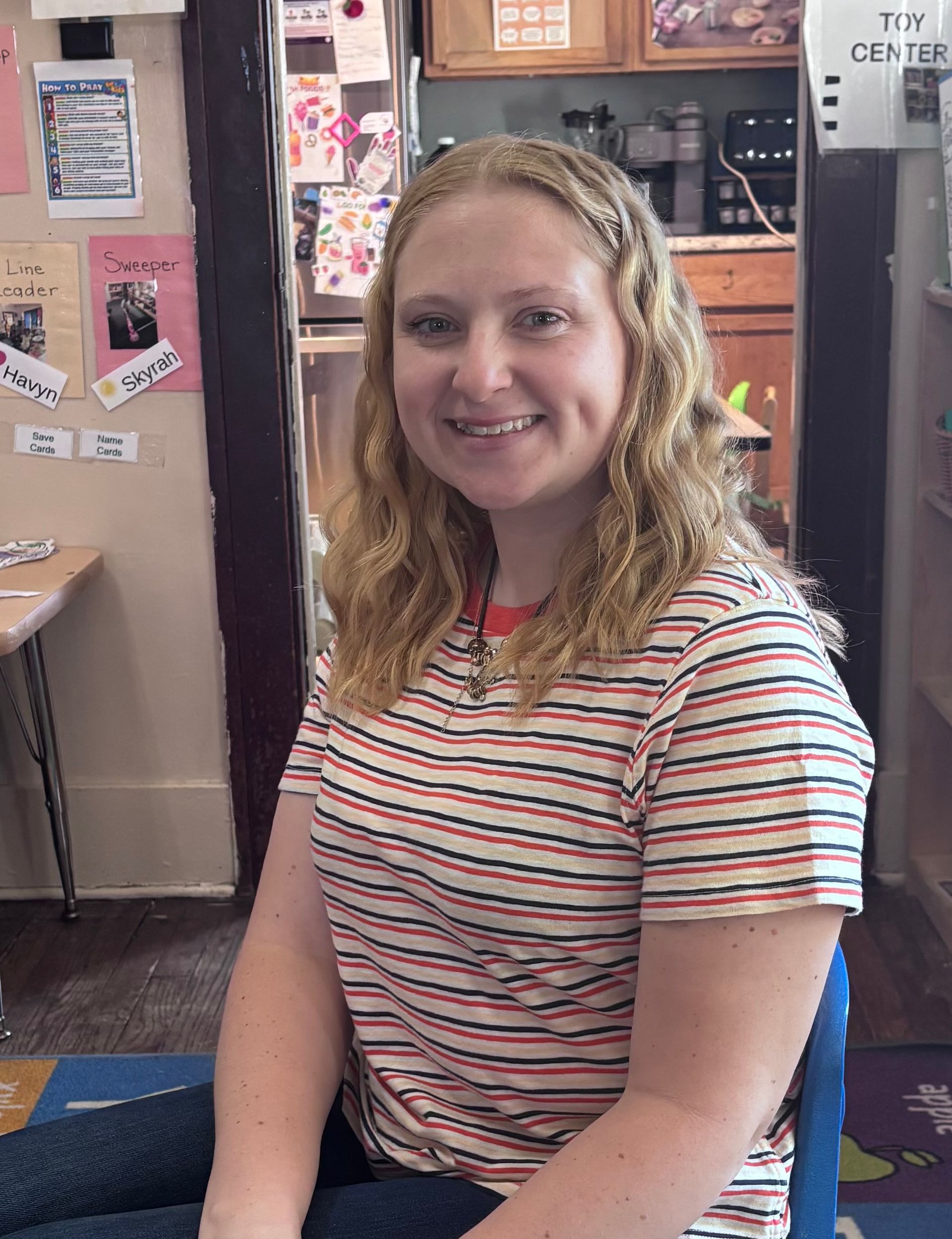Smiling person in a striped shirt seated indoors, with posters and cabinets in the background.