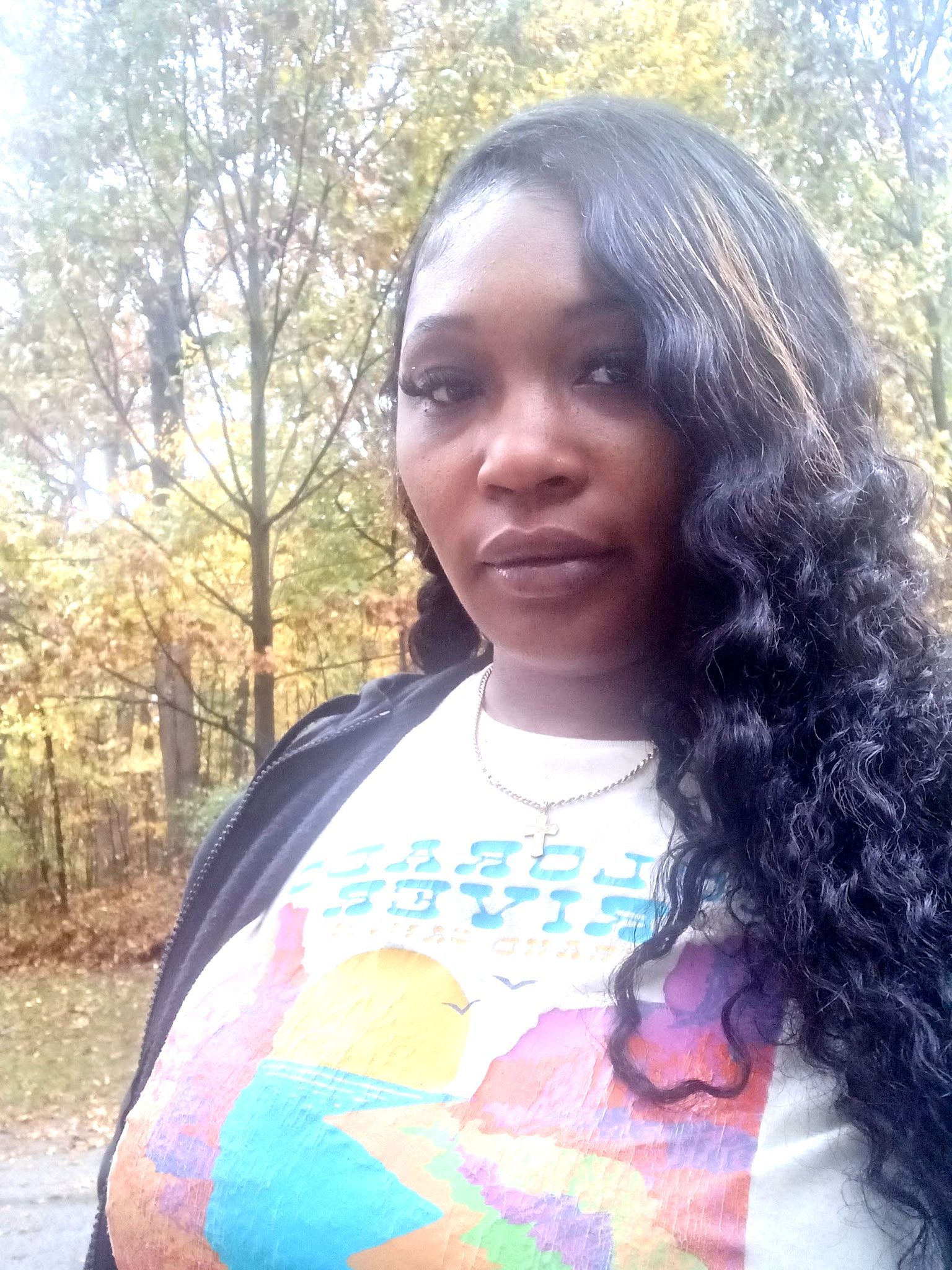 Woman outdoors in a wooded area, wearing a black jacket and colorful graphic T-shirt.