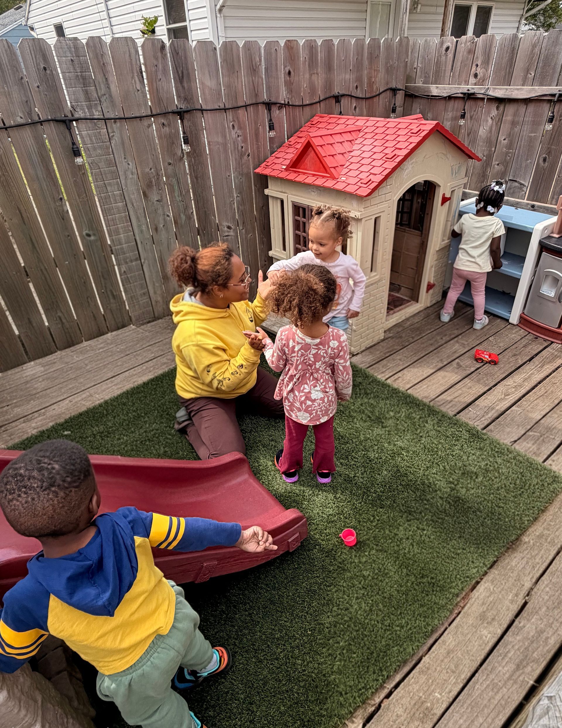 Children playing in a fenced backyard around a red playhouse and slides on turf and wood decking