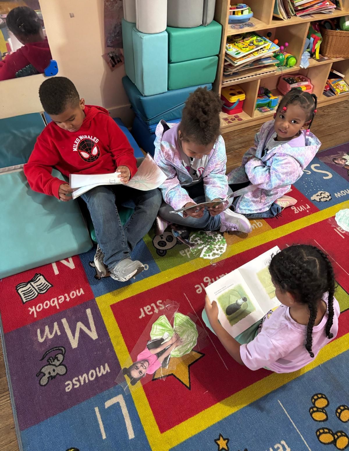 A smiling teacher kneels in a classroom with notebooks as students work at a table in the background.