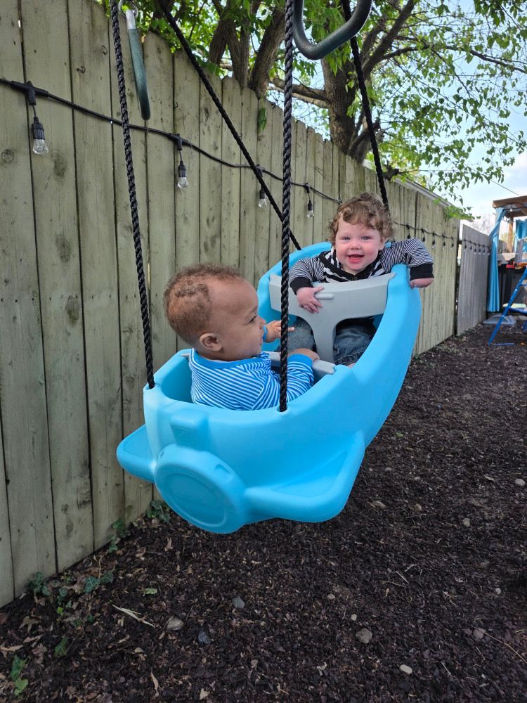 Two children riding in a blue backyard swing by a wooden fence, smiling and facing each other