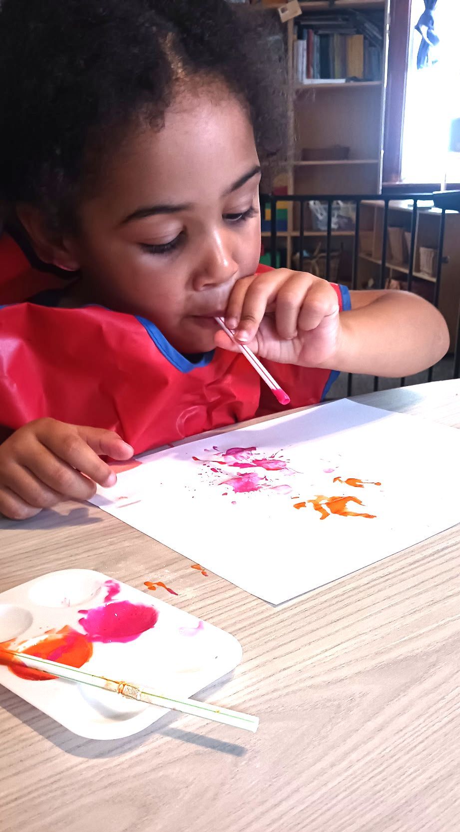 Child painting at a table, concentrating on a red and white art project with a brush.