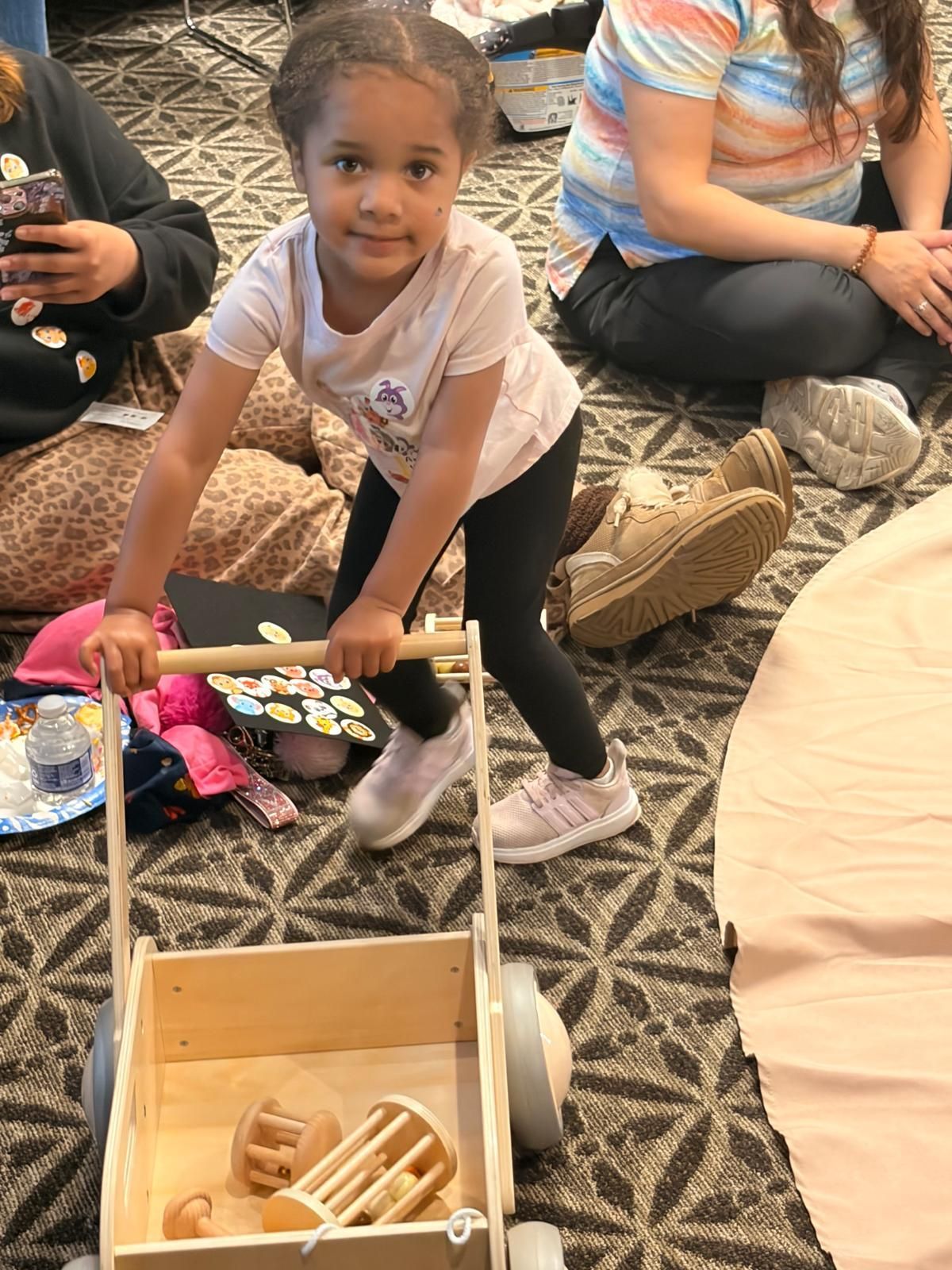 Child kneels on patterned carpet, reaching into a wooden toy bin with a pink toy stethoscope.
