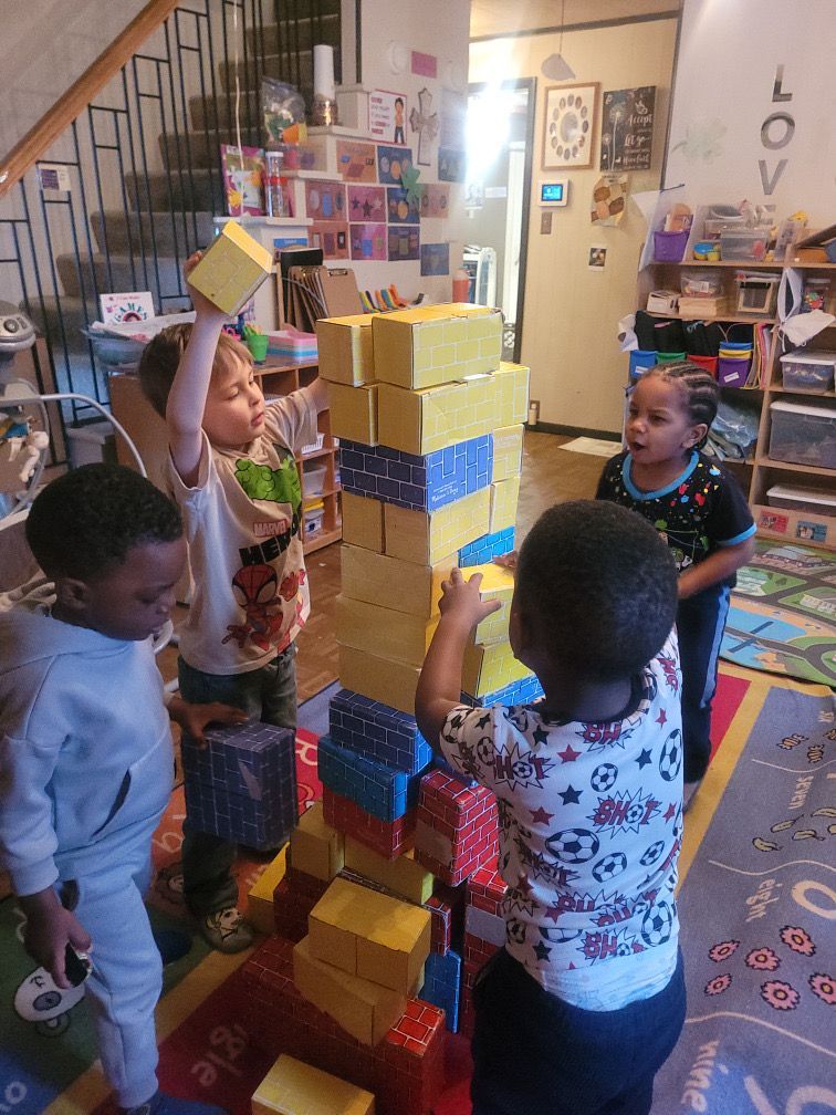 Children stacking colorful blocks into a tall tower in a classroom, with shelves and toys in the background.