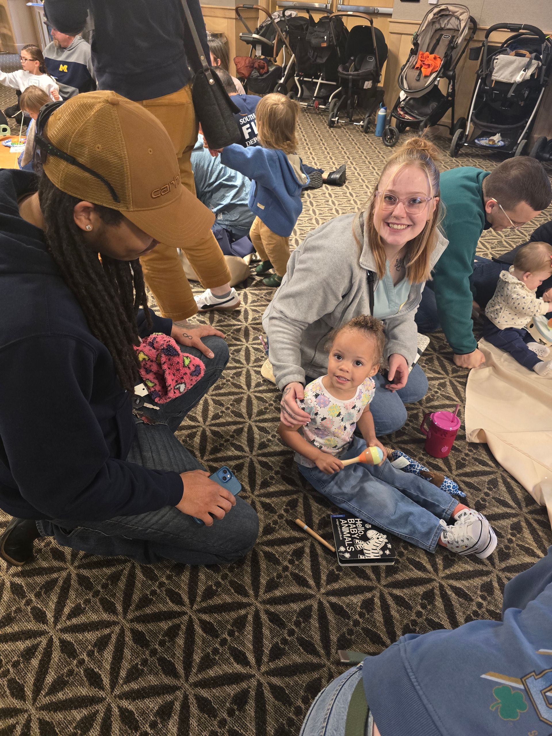 People sitting on a patterned carpet, smiling and interacting in a crowded indoor gathering.