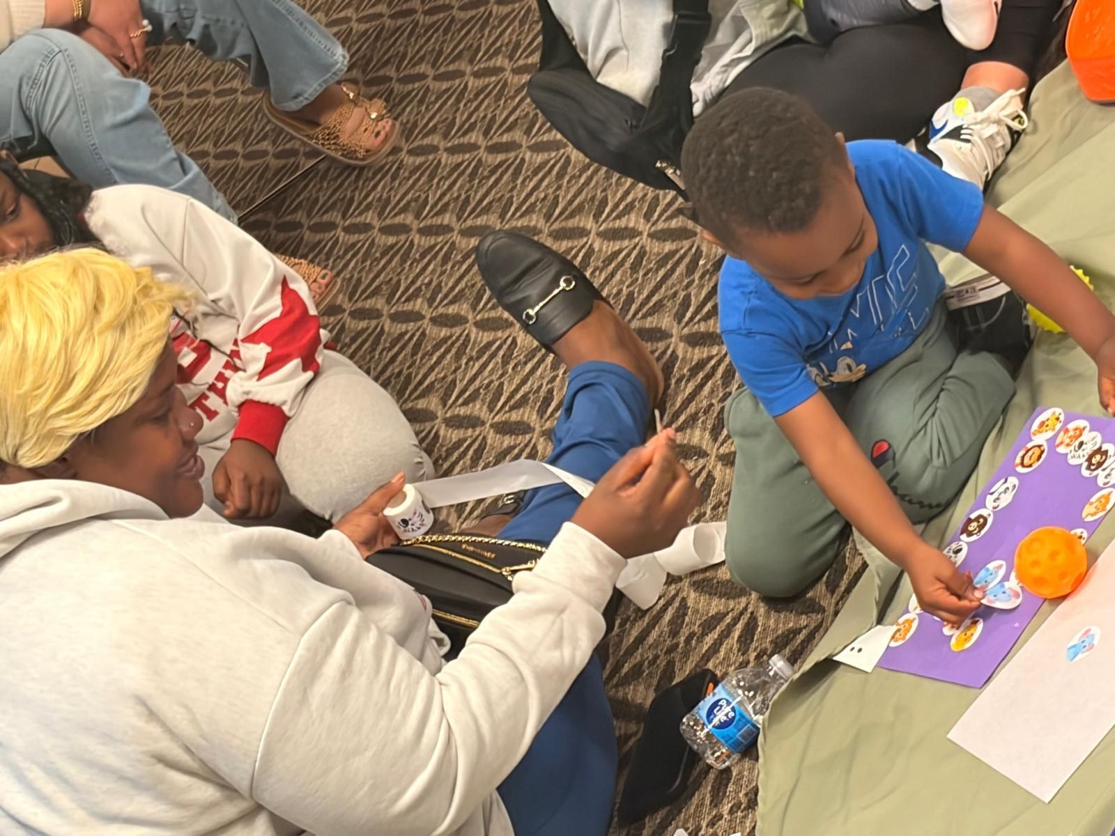 Children sitting on carpet, arranging colorful craft materials on paper during an activity