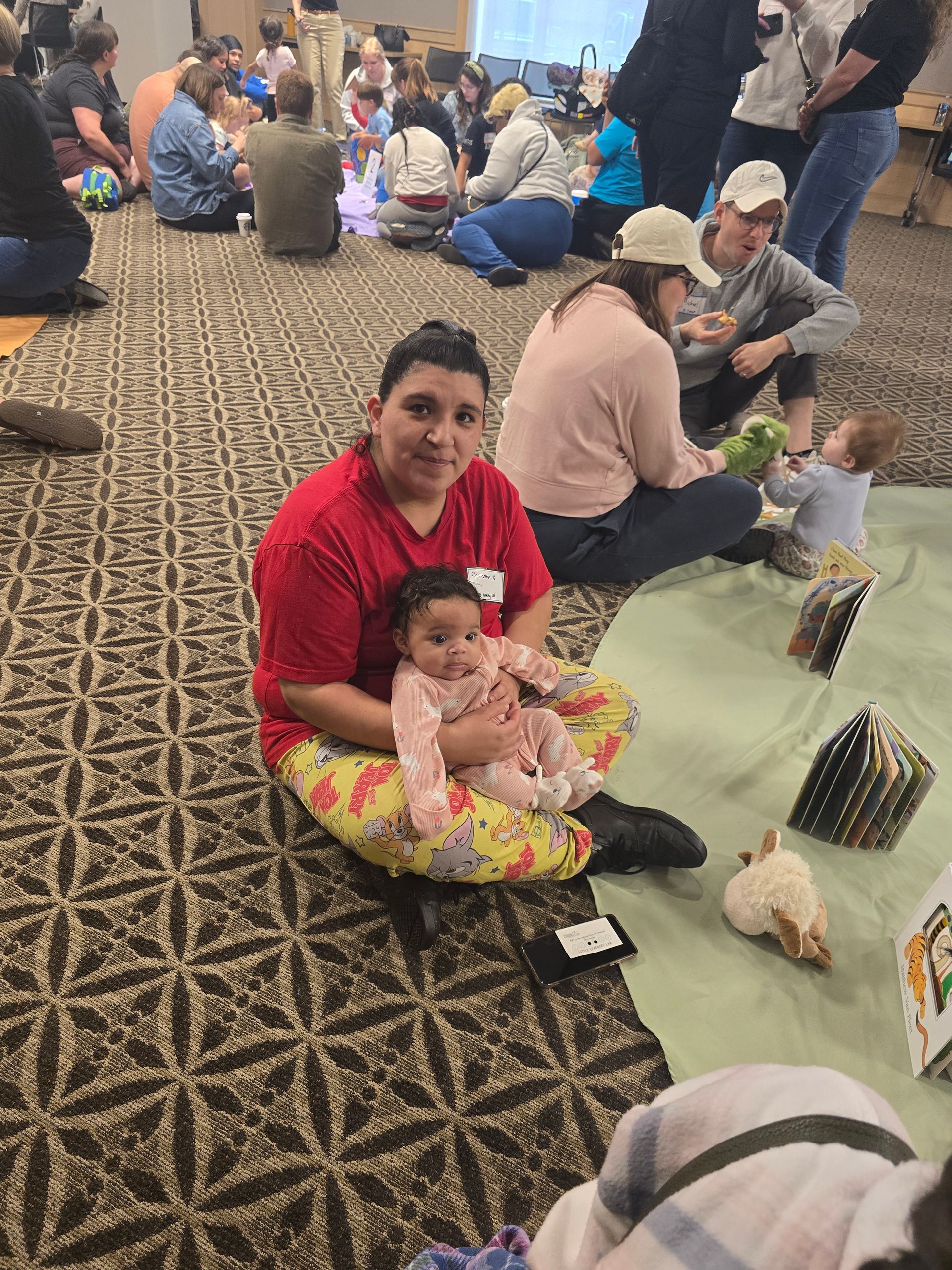 Person in red shirt sits on carpet holding a baby during a crowded indoor gathering