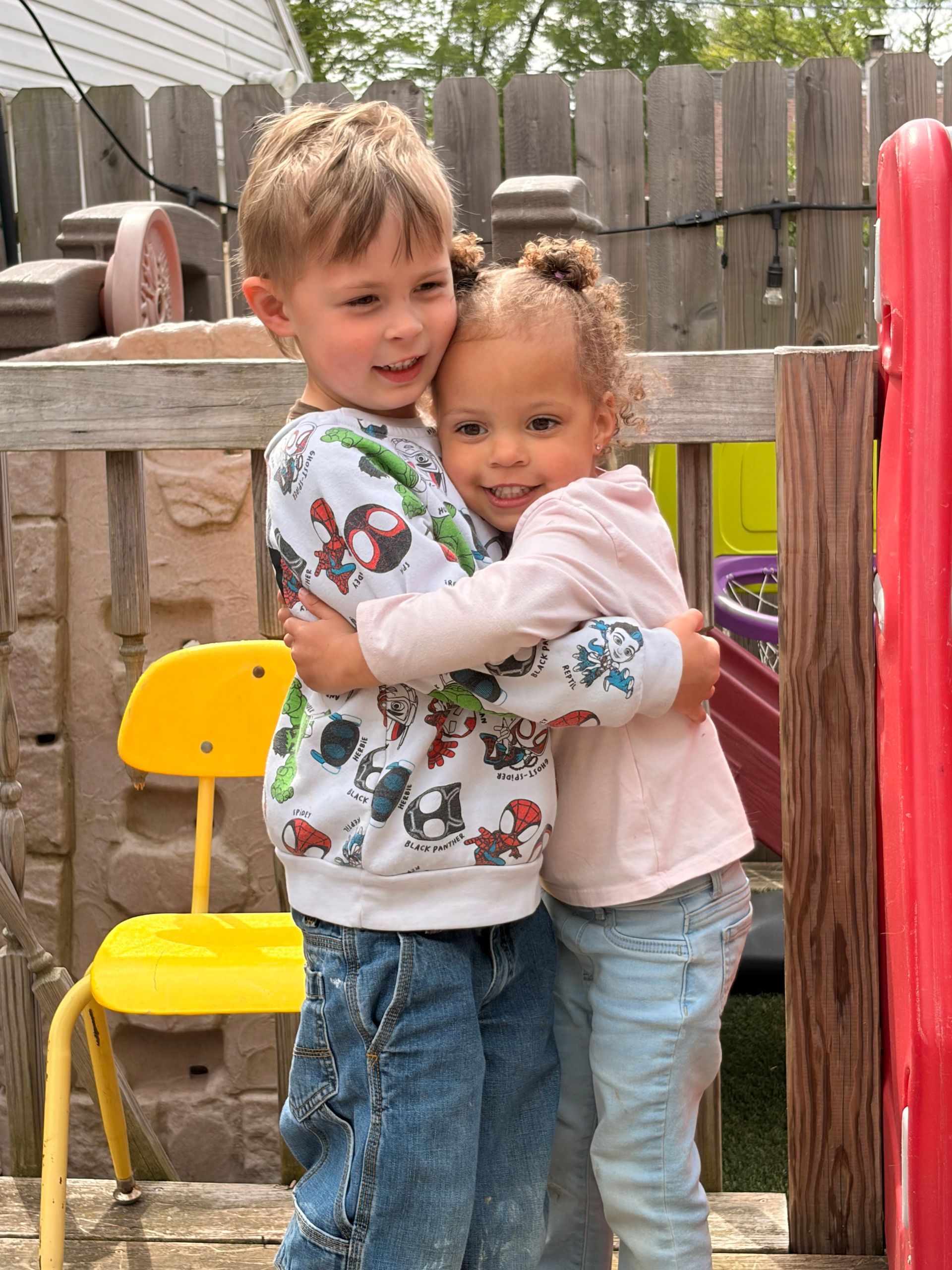 Two children hugging outdoors near a yellow chair and wooden fence