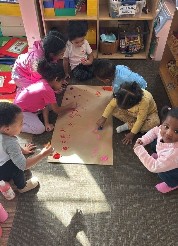 Children gathered around paper on the floor, painting with red dots in a classroom.