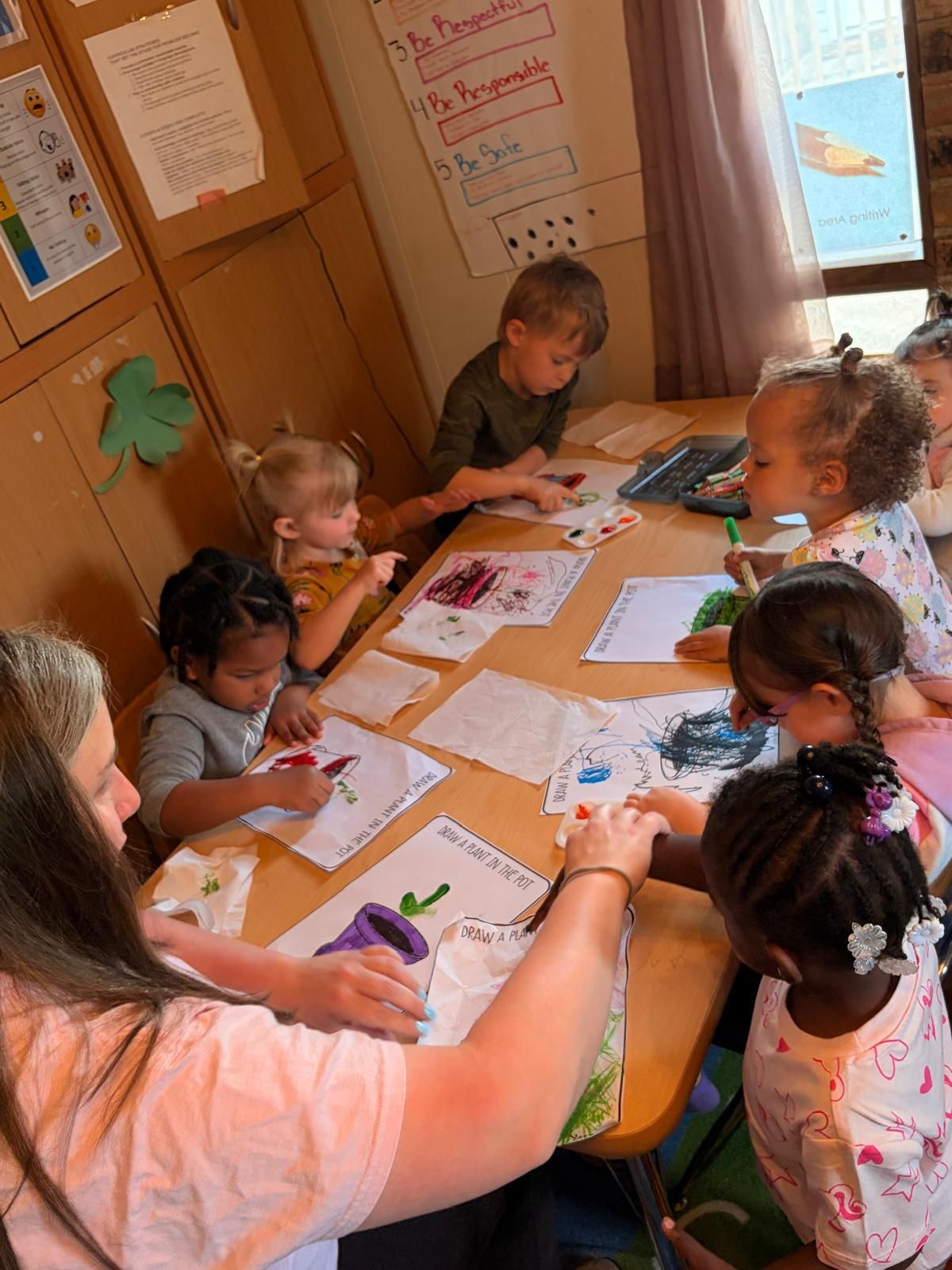 Children and a teacher gather around a table, coloring and drawing in a small classroom.