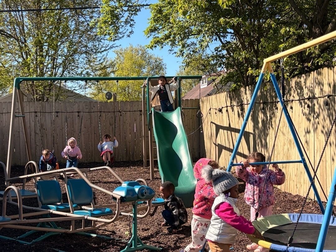 Children play on a backyard slide and trampoline under a wooden fence and trees.