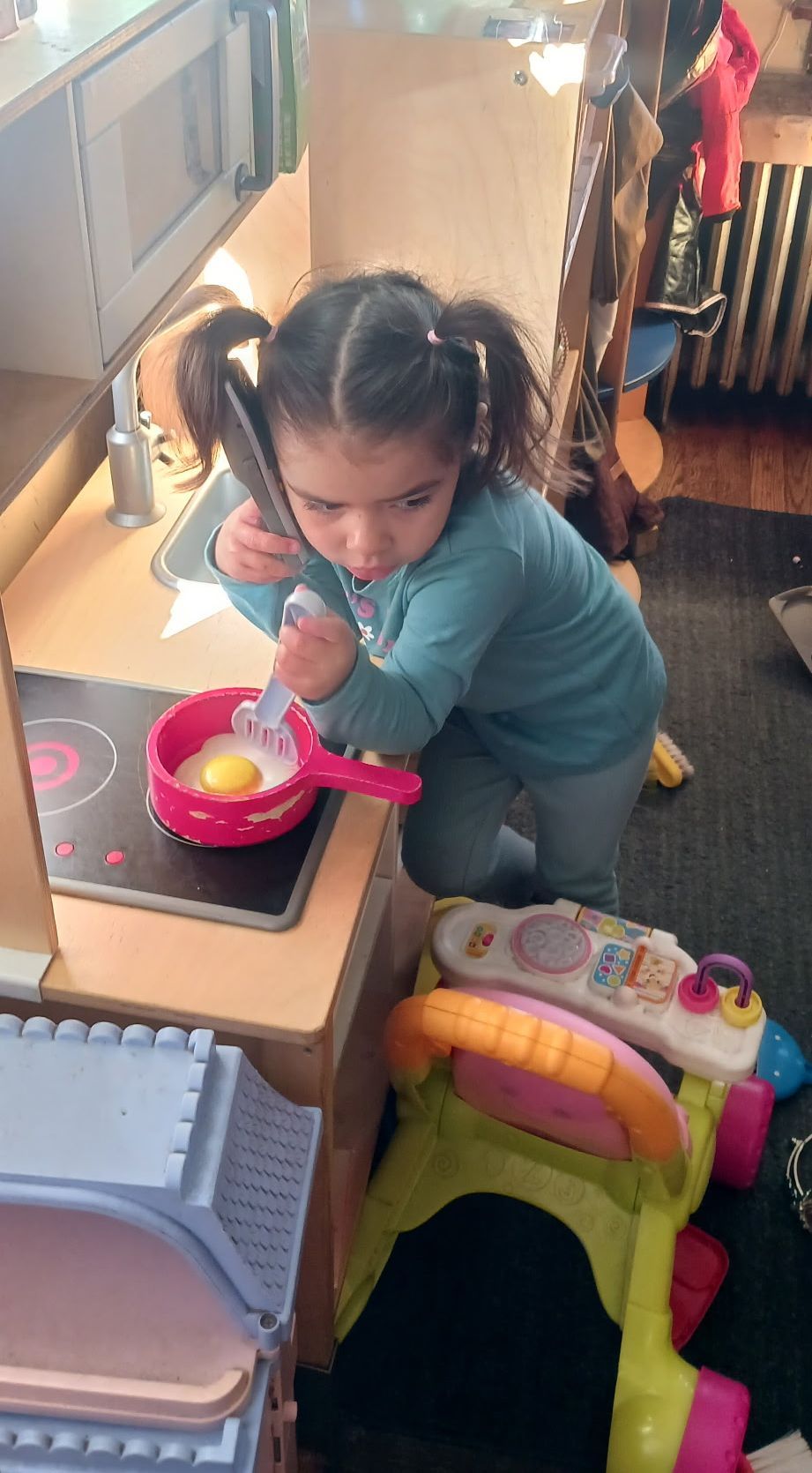 Child playing with a toy kitchen set indoors, leaning over a pink play stove.