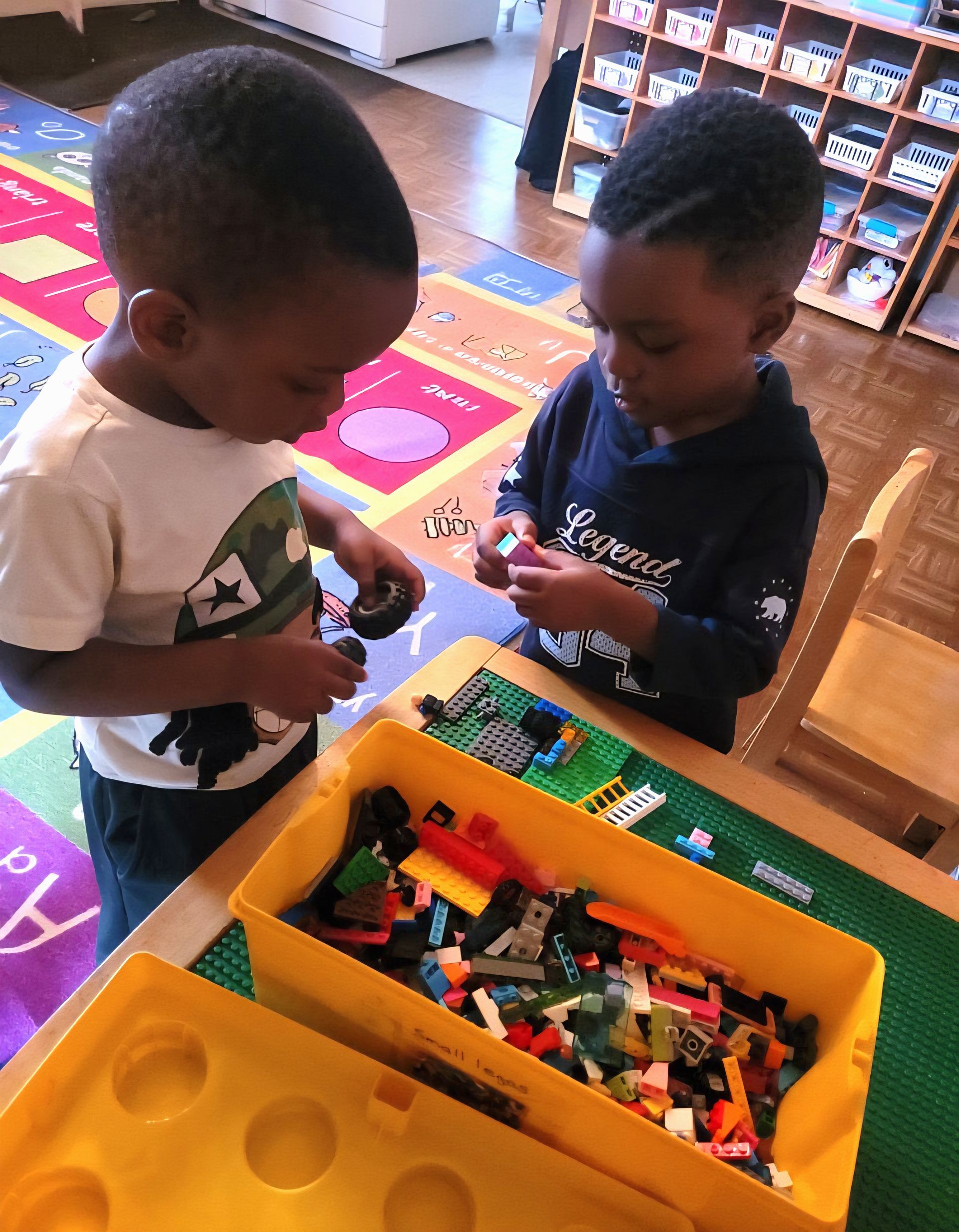 Two children standing by a bin of LEGO bricks, building together on a colorful classroom rug.