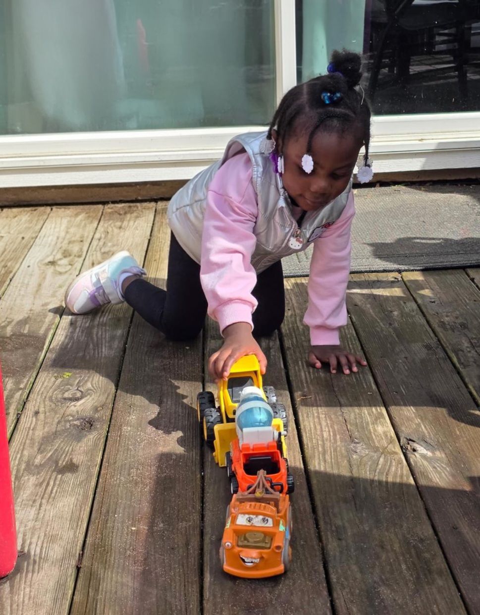 Child kneeling on a wooden deck, pushing toy trucks in a row toward the camera.