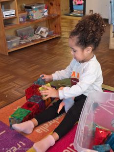 A teacher sits on a colorful floor mat, joyfully hugging two students in a bright classroom filled with learning toys.