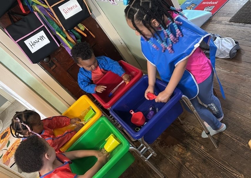 Children in colorful bins, each looking down at small items in a classroom activity area.
