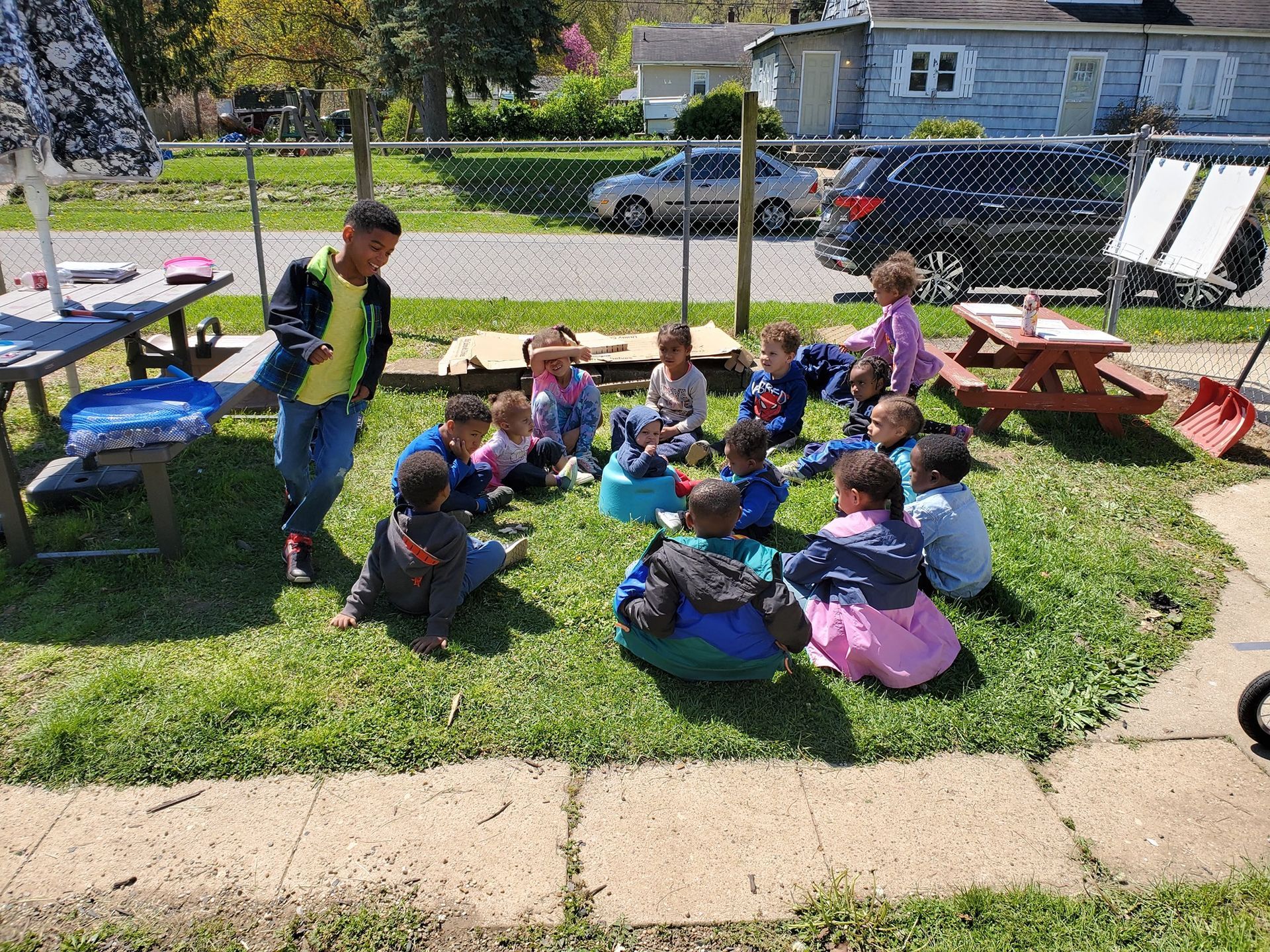 Children sitting on blankets in a sunny park, with picnic tables and a chain-link fence behind them.
