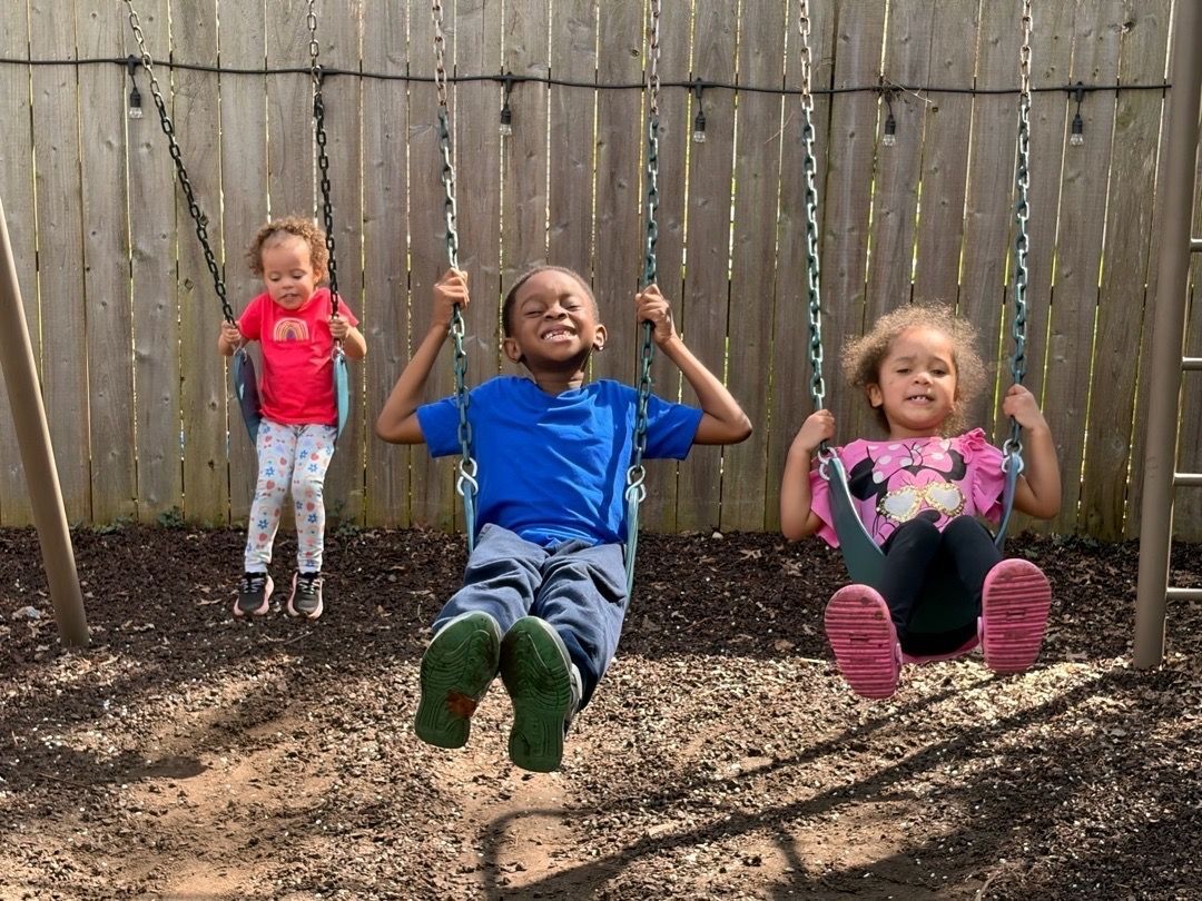 Three children on backyard swings in front of a wooden fence, smiling and mid-swing.