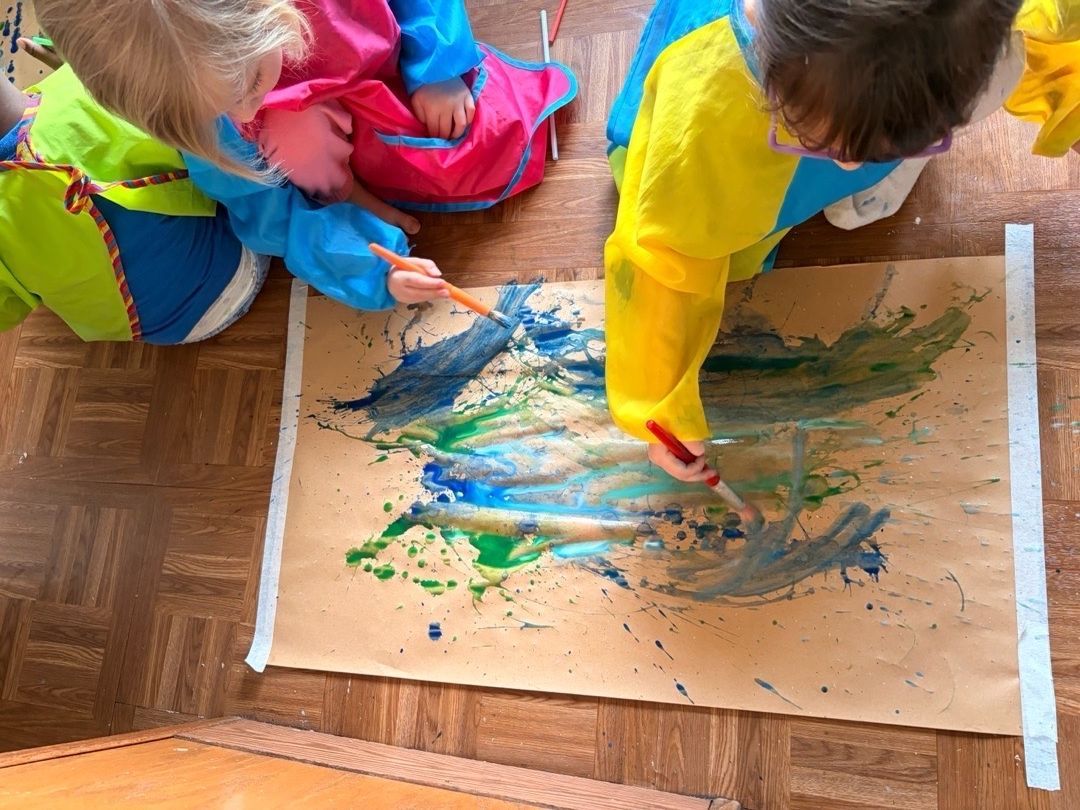 Children painting with blue, green, and yellow paint on a large sheet of paper on the floor